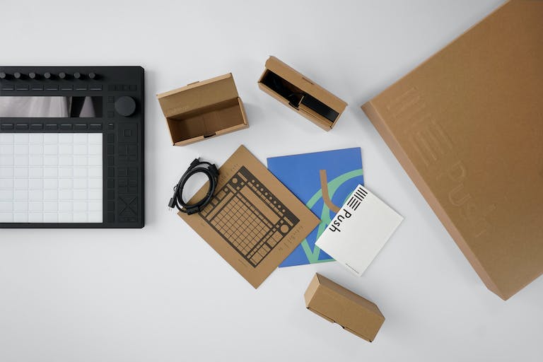 Push being unboxed, shot from above. Push is off to the left side, with brown cardboard packaging, an onboarding guide, folded poster and USB-C cable laid out on the desk to its right.